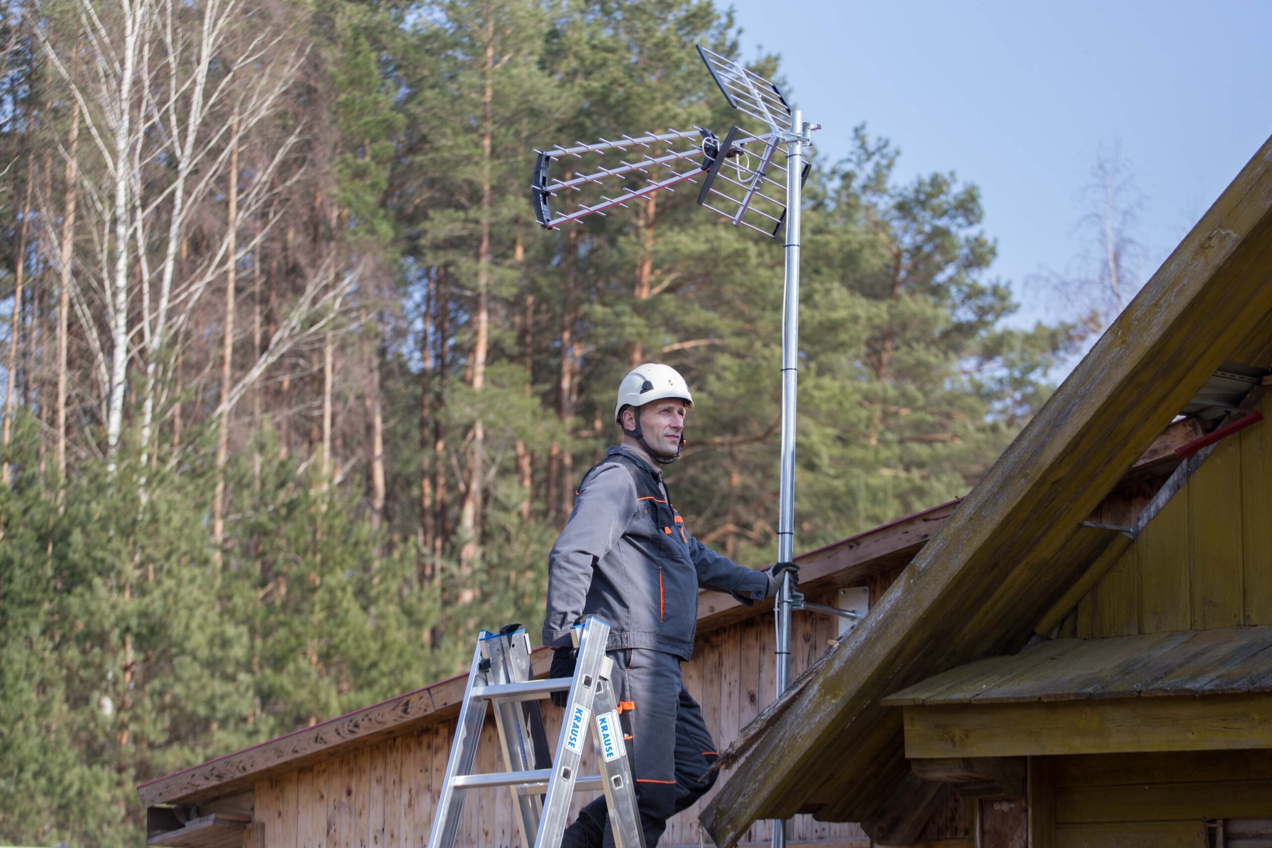 Telecentro inžinierius ant kopėčių matuoja TV signalo lygį ties namų stogu